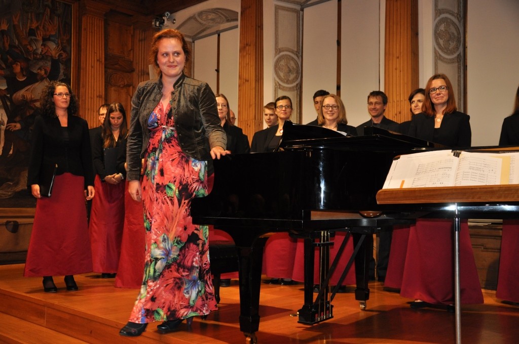 Eine Sängerin steht am Klavier, der Chor im Hintergrund.